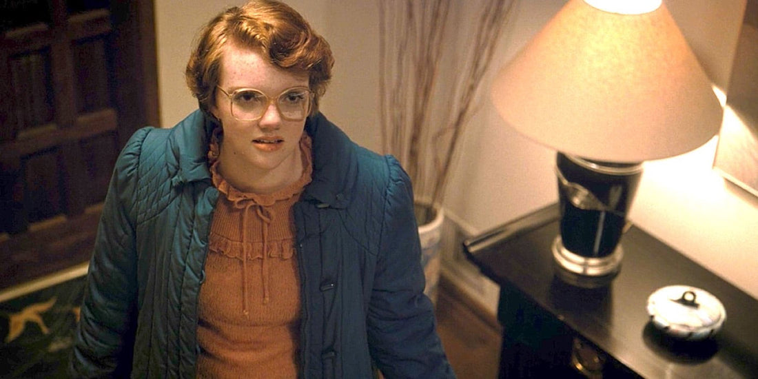 Shannon Purser as Barbara Holland, glasses and a blue coat, in a hallway.
