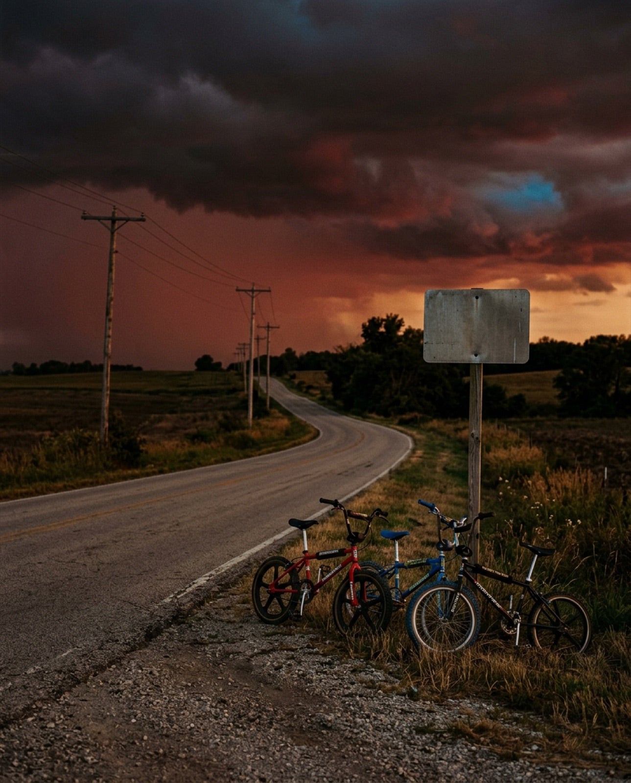 Three BMX bikes at the edge of an American 80s country road, blank sign and power poles under a red storm sky